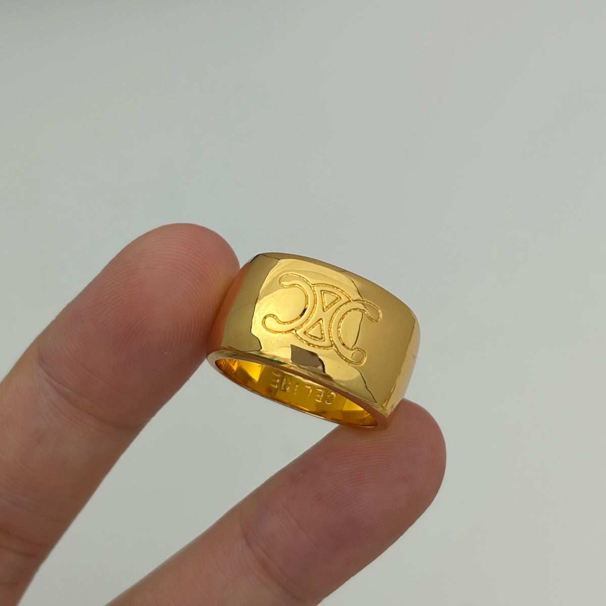 A close-up of a gold ring with an engraved interlocking CC monogram, held between two fingers against a plain light gray background.