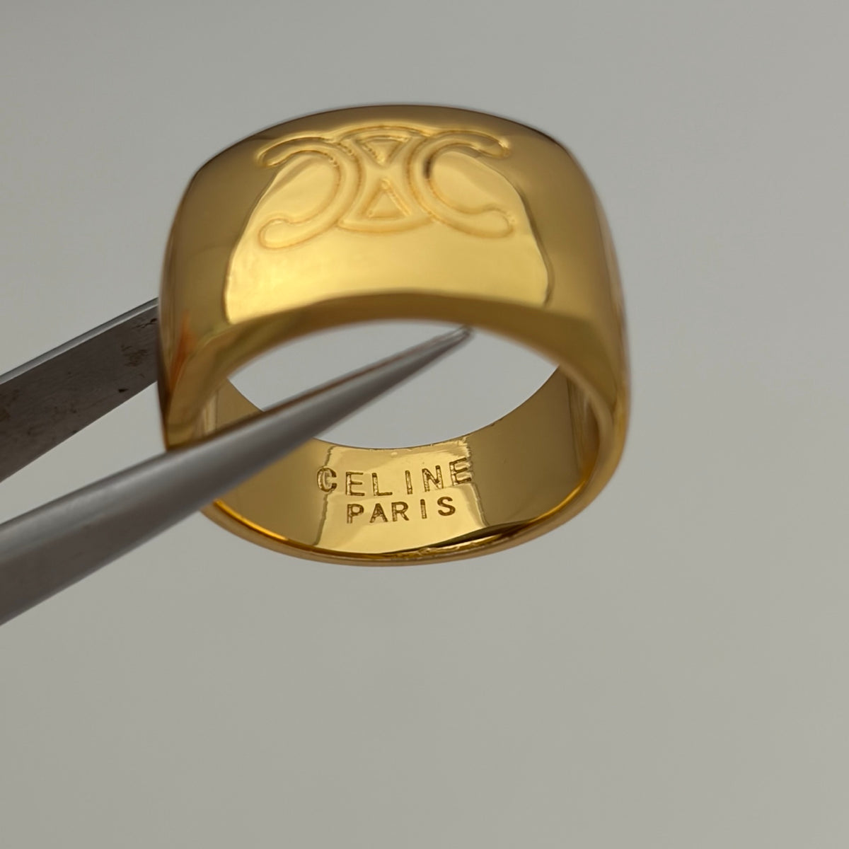 A gold Celine ring with a prominent CC logo on the exterior and 'CELINE PARIS' stamped on the interior, held by metal tweezers against a neutral gray background.