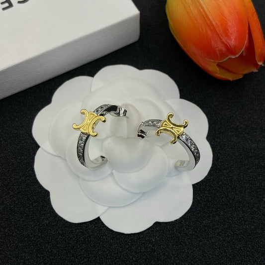 A pair of silver-toned hoop earrings with gold-toned Celine Triomphe logos and a row of clear crystals, displayed on a white flower-shaped stand.