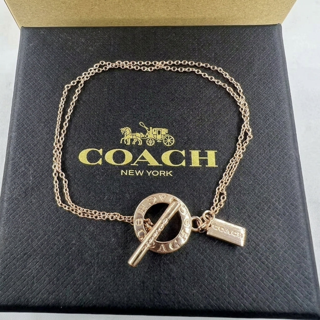 A gold-tone Coach New York chain bracelet with a circular toggle clasp and a small rectangular tag, displayed on top of a black Coach New York box.