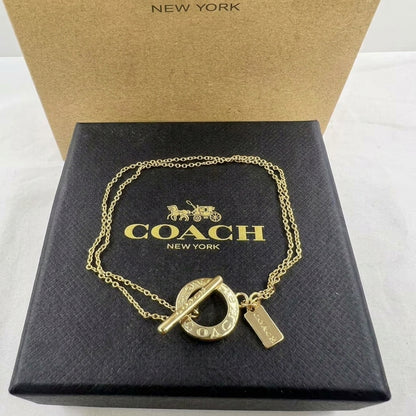A gold-tone Coach New York toggle ring necklace with a signature carriage logo charm and a small rectangular tag, displayed on a black textured Coach New York box with its kraft paper box behind it.