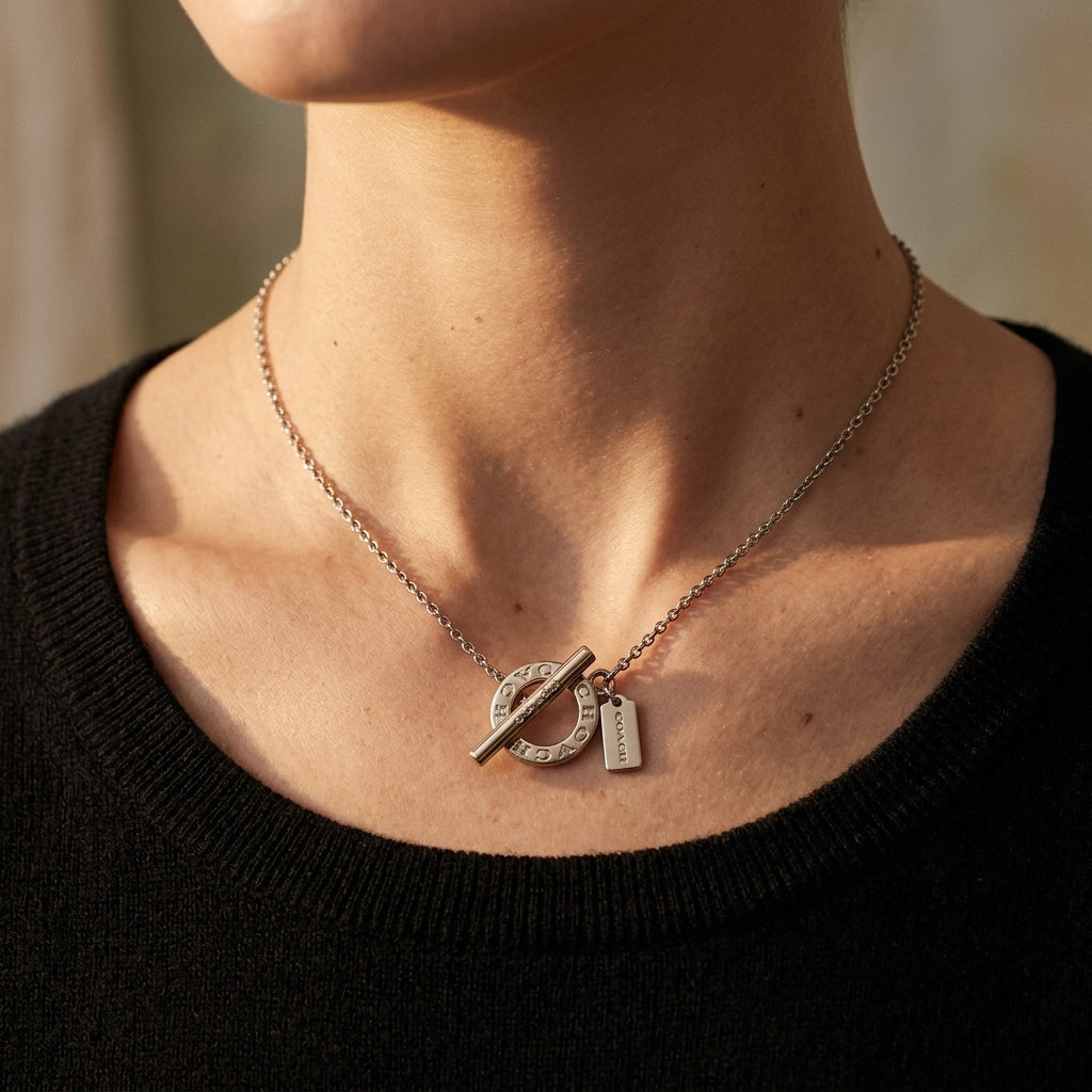 A close-up of a person wearing a delicate silver chain necklace with a toggle-style pendant and a small rectangular tag, shown against the neck and upper chest.