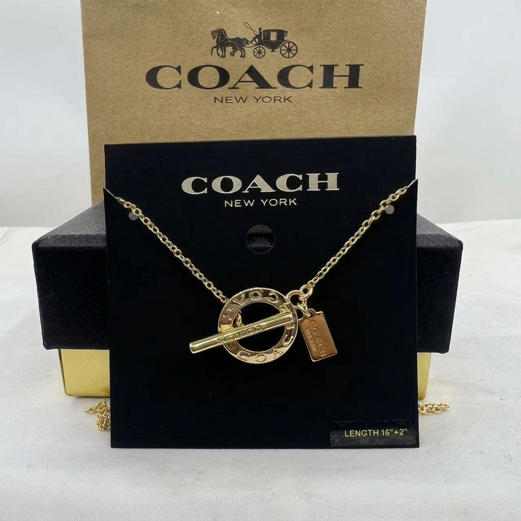 A gold-tone Coach necklace with a circular ring charm, toggle bar, and small rectangular tag, displayed on a black Coach New York card with a kraft Coach box behind it.