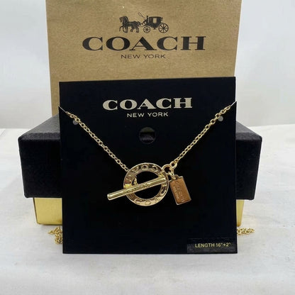 A gold-tone Coach necklace with a circular ring charm, toggle bar, and small rectangular tag, displayed on a black Coach New York card with a kraft Coach box behind it.