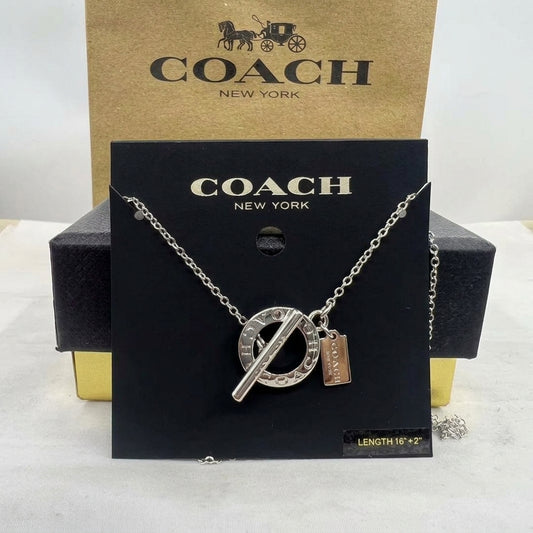 A Coach New York silver-tone necklace with a circular interlocking O pendant and a small rectangular tag charm, displayed on a black branded card in front of a Coach New York box.