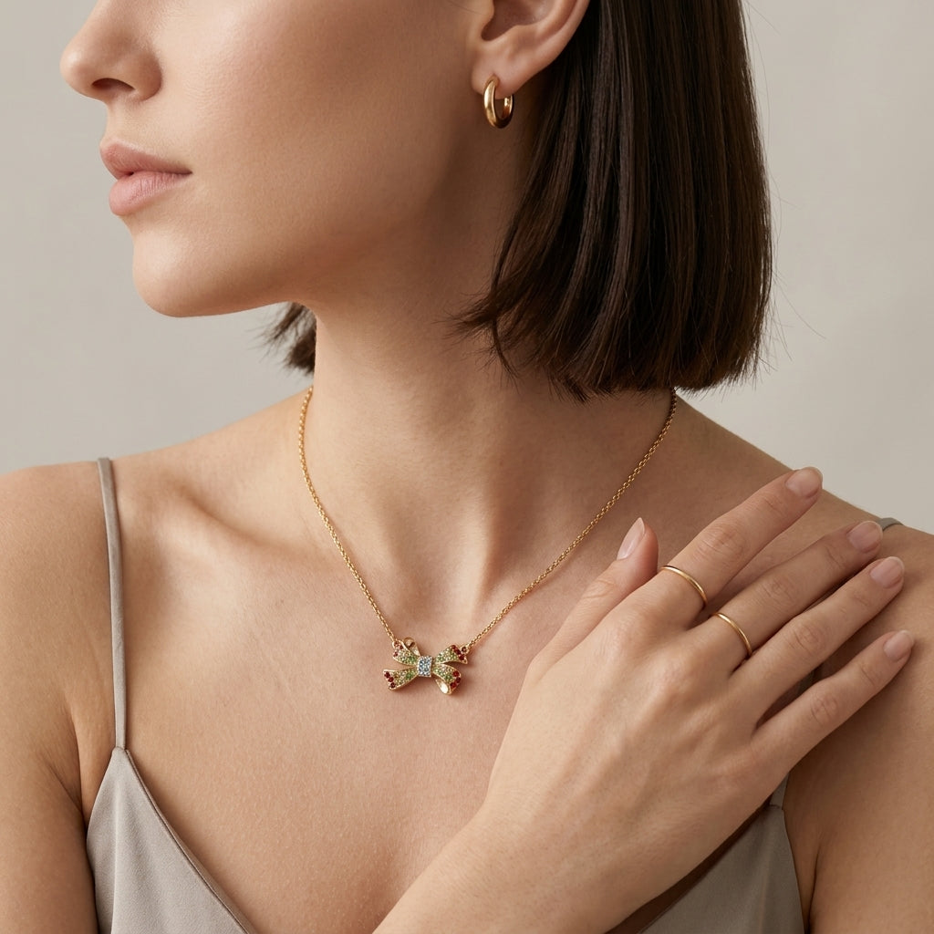 A close-up lifestyle shot of a woman wearing a delicate gold necklace with a small bow-shaped pendant adorned with green and pink stones, along with matching gold hoop earrings and rings.