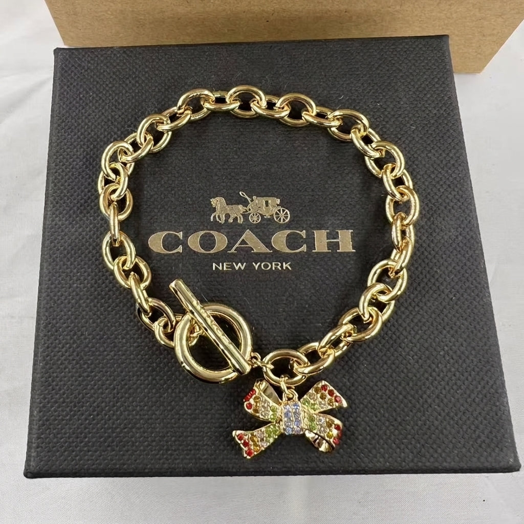 A gold-tone Coach New York chain bracelet with a toggle clasp and a bow charm embellished with red, green, and white rhinestones, displayed on its black textured Coach New York box.