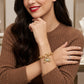 A woman wearing a gold butterfly bracelet with a small green gemstone and a matching gold ring, styled with a brown ribbed knit sweater.
