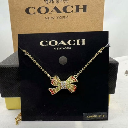 A Coach New York necklace displayed on a black branded card, featuring a gold-tone chain and a bow-shaped pendant with red and yellow rhinestone accents and a clear center stone. A tan Coach box is visible behind the card.