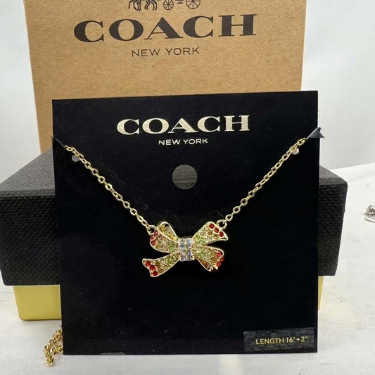 A Coach New York necklace displayed on a black branded card, featuring a gold-tone chain and a bow-shaped pendant with red and yellow rhinestone accents and a clear center stone. A tan Coach box is visible behind the card.
