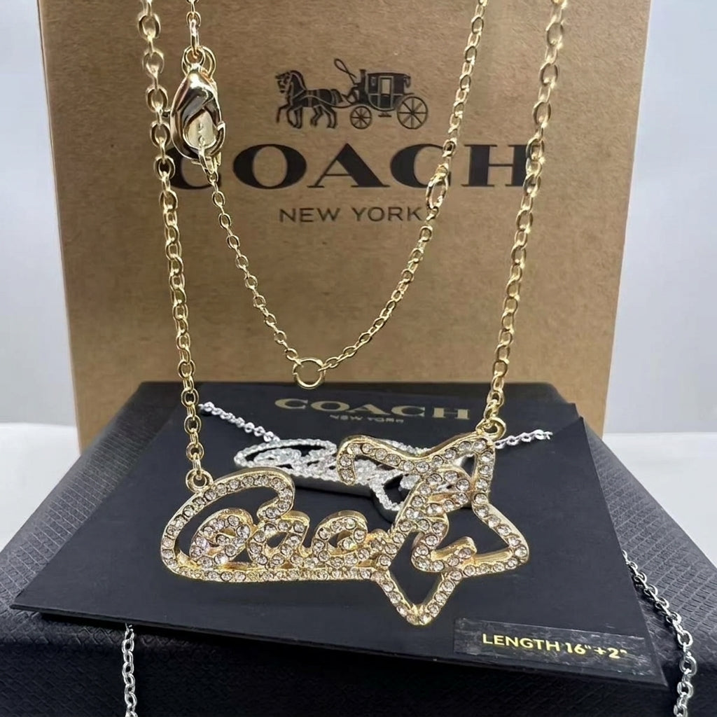 A gold-tone Coach name necklace with a star-shaped pendant set with clear rhinestones, displayed on a black Coach presentation card in front of a brown Coach box.