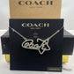 A silver-tone Coach script necklace paved with clear rhinestones, displayed on its black branded card packaging with a brown Coach box behind it.