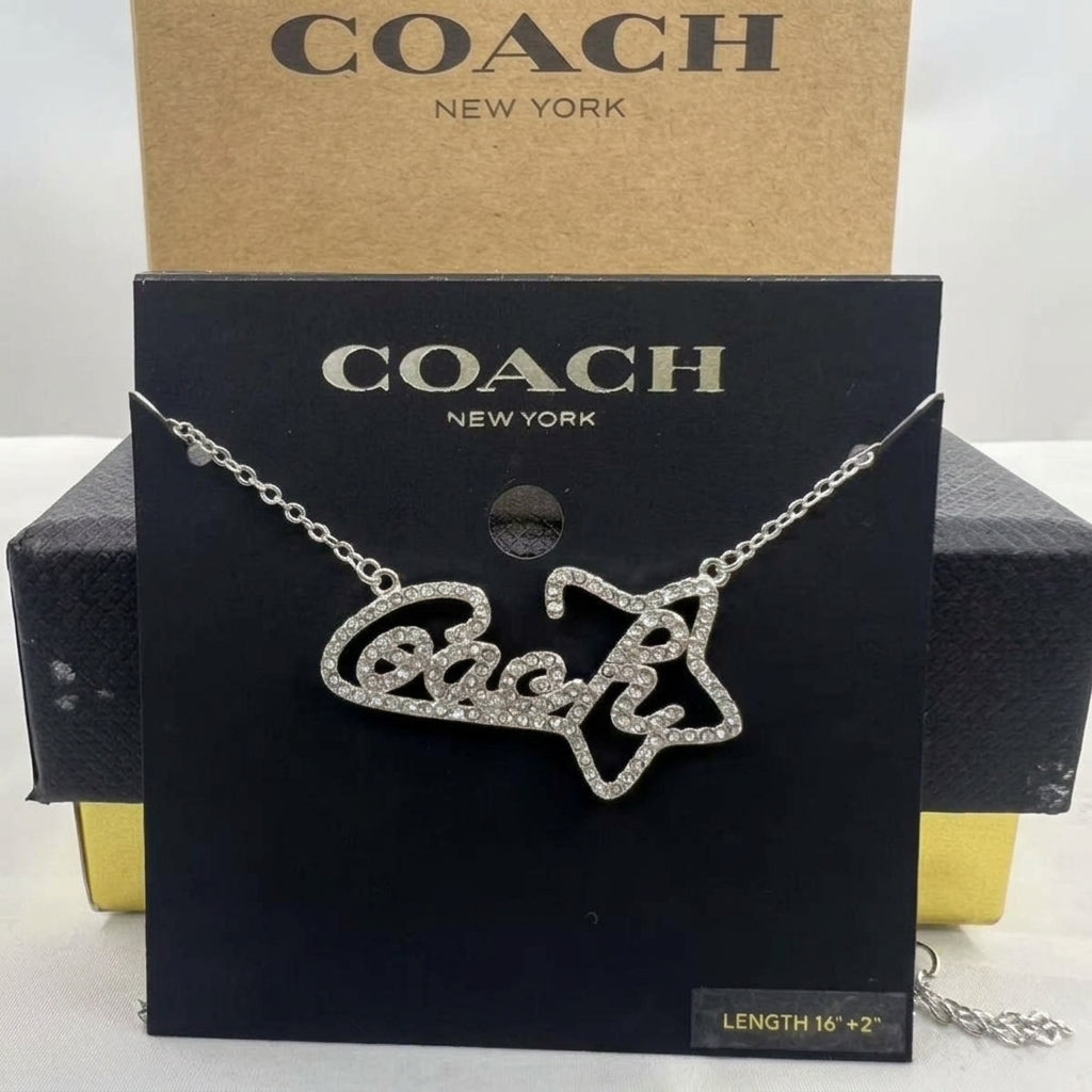 A silver-tone Coach script necklace paved with clear rhinestones, displayed on its black branded card packaging with a brown Coach box behind it.