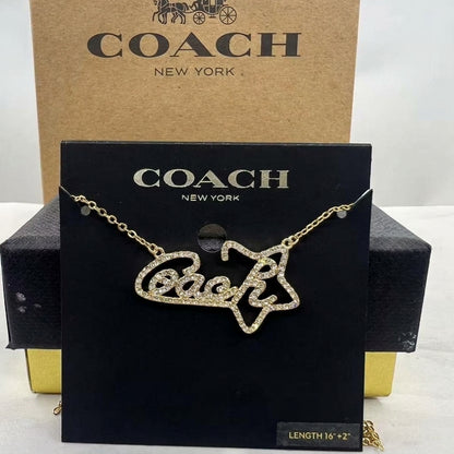 A gold-tone Coach necklace with a star-shaped pendant featuring the word 'Coach' in rhinestones, displayed on a black branded card with a brown Coach box behind it.