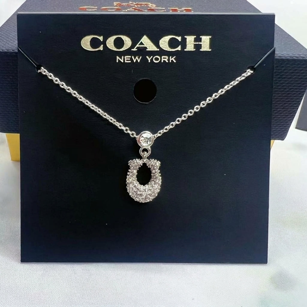 A silver-tone Coach New York necklace with a horseshoe-shaped pendant encrusted with clear stones, displayed on a black Coach New York branded card.