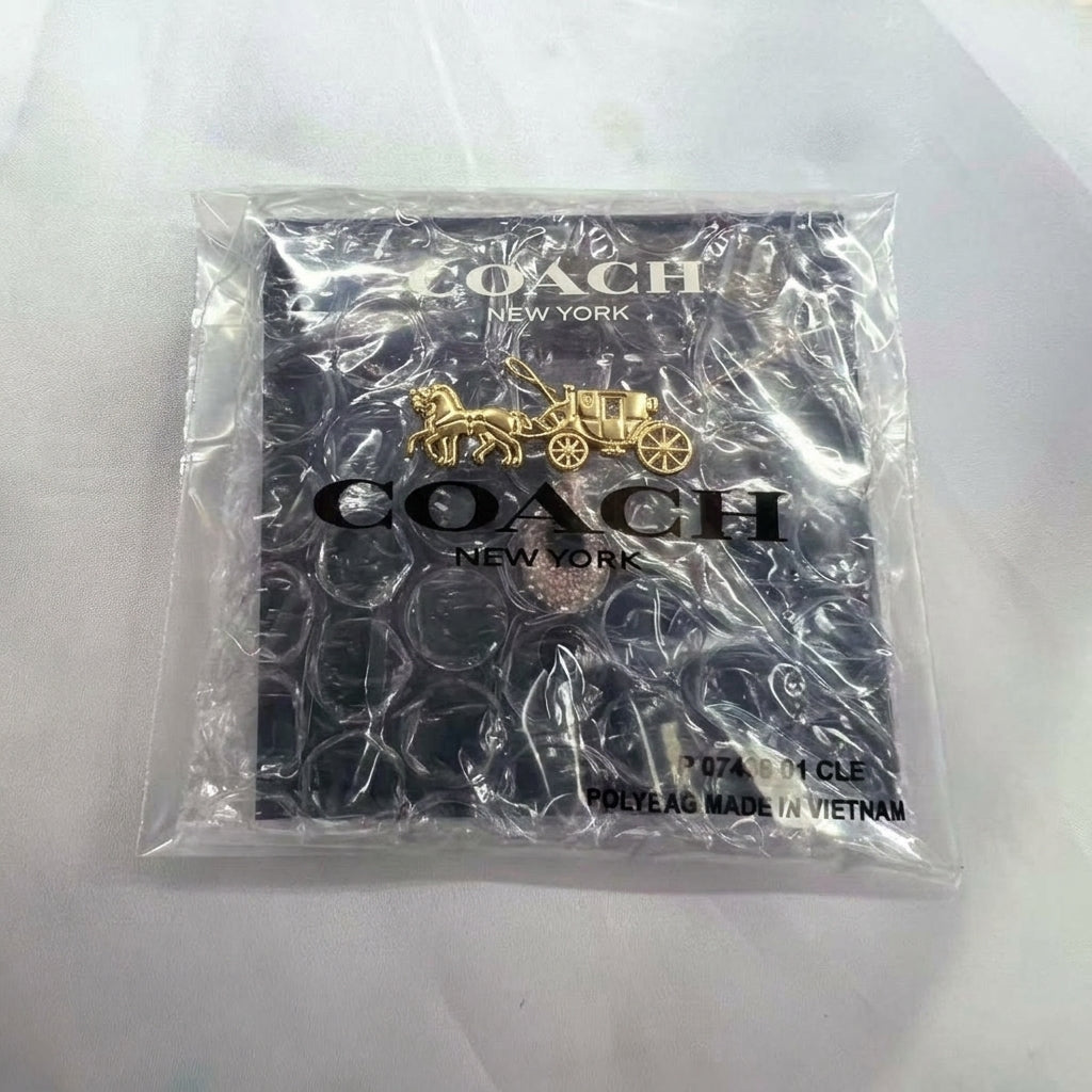 A Coach New York logo keychain in a gold-tone horse-drawn carriage design, packaged inside a clear bubble bag with a black branded backing card on a white background.