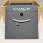 A Coach New York necklace displayed on a branded dark gray card, with a rose-gold-toned chain, a curved bar set with small clear stones, and a dangling letter C charm. The card is placed in front of an open gold-tone box.