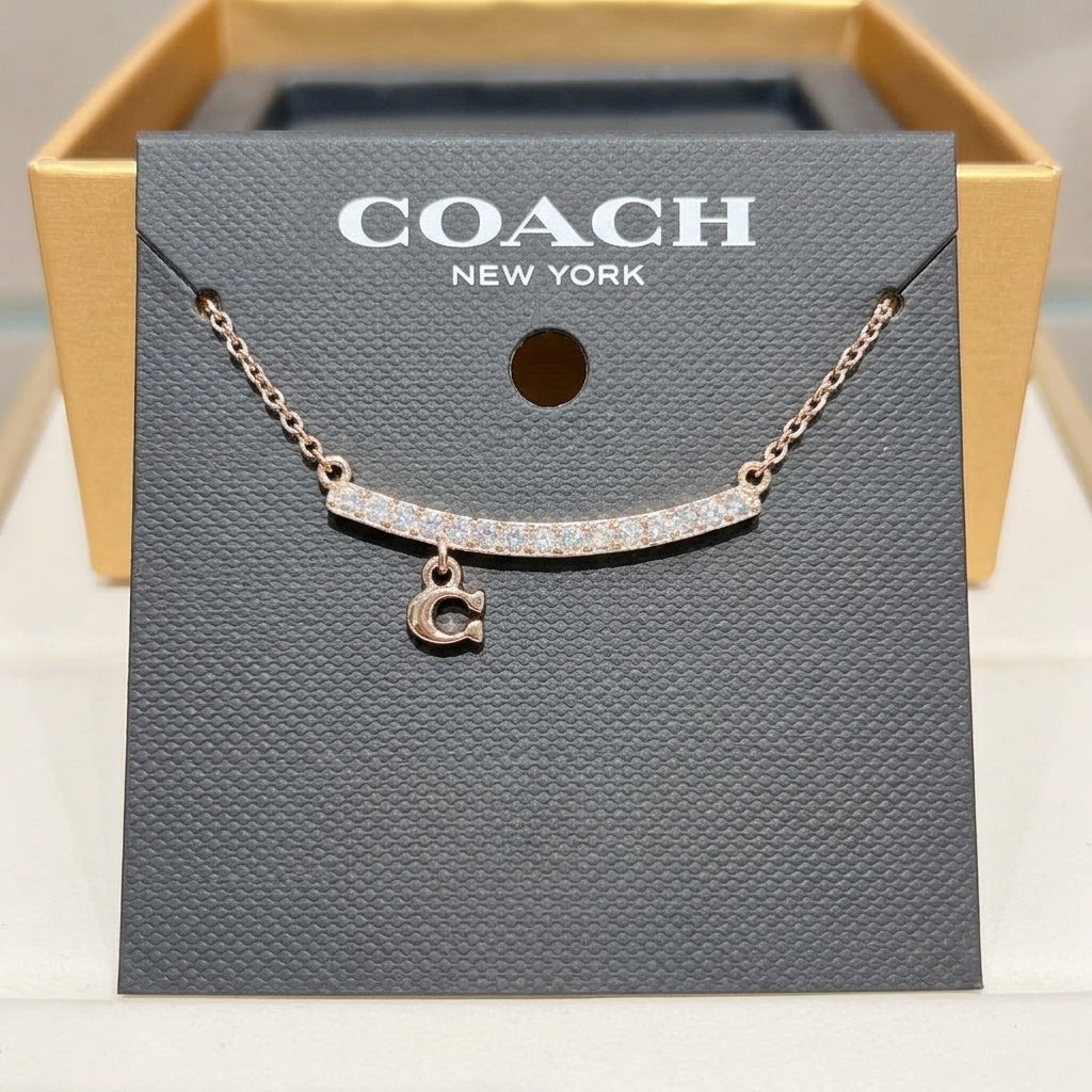 A Coach New York necklace displayed on a branded dark gray card, with a rose-gold-toned chain, a curved bar set with small clear stones, and a dangling letter C charm. The card is placed in front of an open gold-tone box.