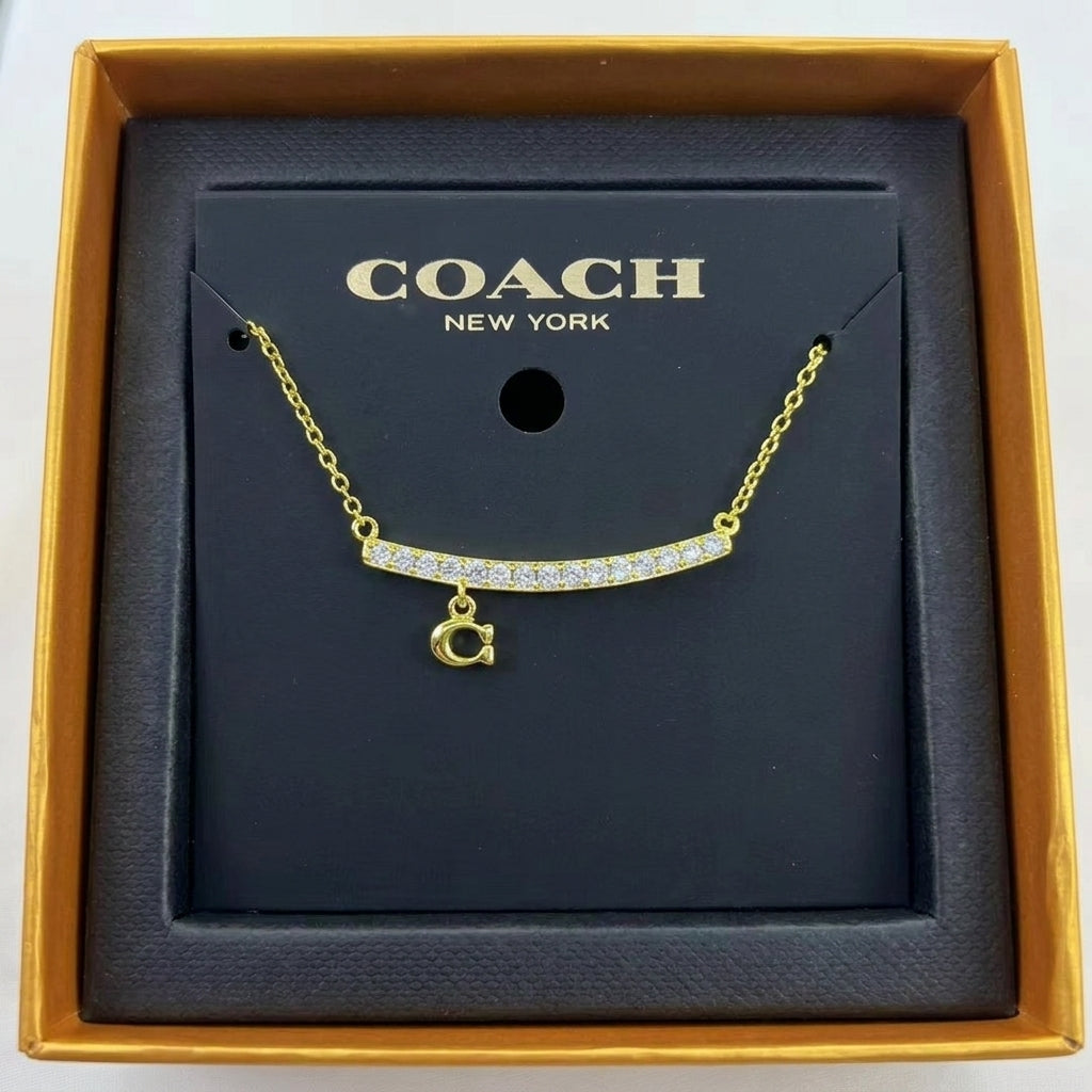 A gold-tone Coach necklace with a curved bar set with clear stones and a dangling 'C' initial charm, presented on a black Coach New York display card inside an open gold box.