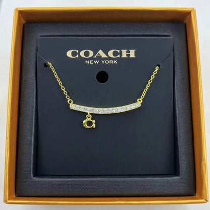A gold-tone Coach necklace with a curved bar set with clear stones and a dangling 'C' initial charm, presented on a black Coach New York display card inside an open gold box.