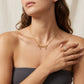 A close-up lifestyle shot of a model wearing a gold script nameplate necklace that spells 'Coach' with three small dangling heart charms, paired with a matching gold ring.