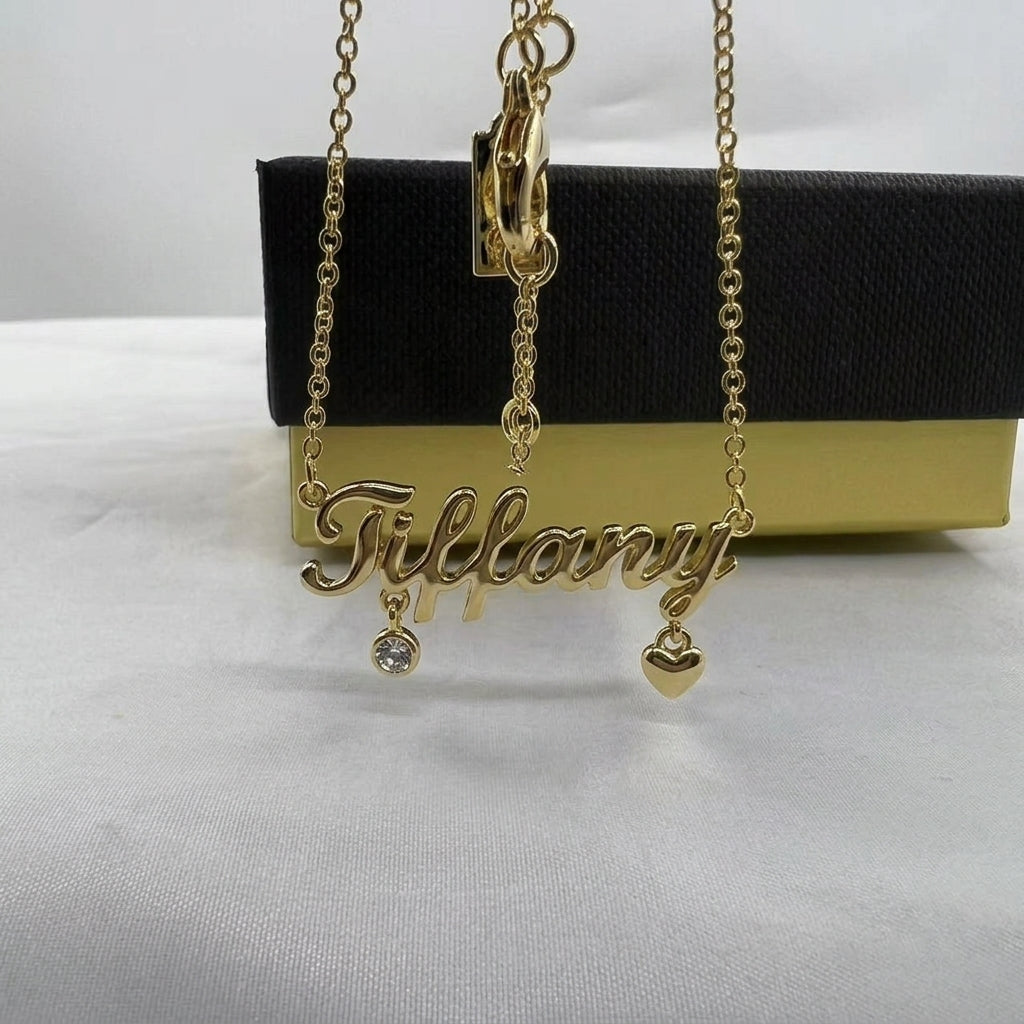 A gold nameplate necklace spelling 'Tiffany' with a small clear crystal charm and a gold heart charm, displayed in front of a black and gold Tiffany box.