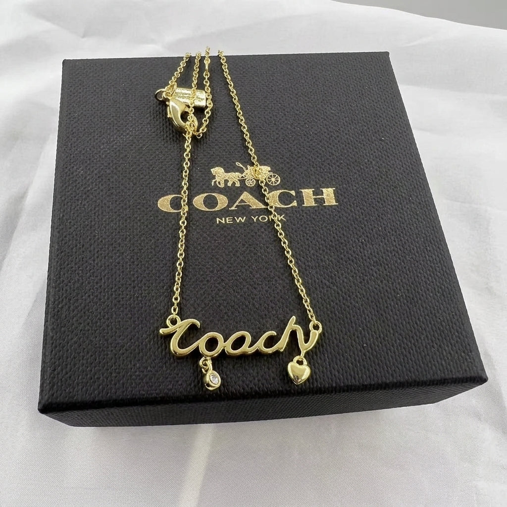 A gold-tone Coach name necklace with a 'Coach' script pendant, a small teardrop charm, and a heart charm, displayed on top of its black branded box.