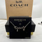 A silver-tone Coach nameplate necklace with a dangling heart charm and clear rhinestone, displayed on a black Coach box against a brown Coach New York box.