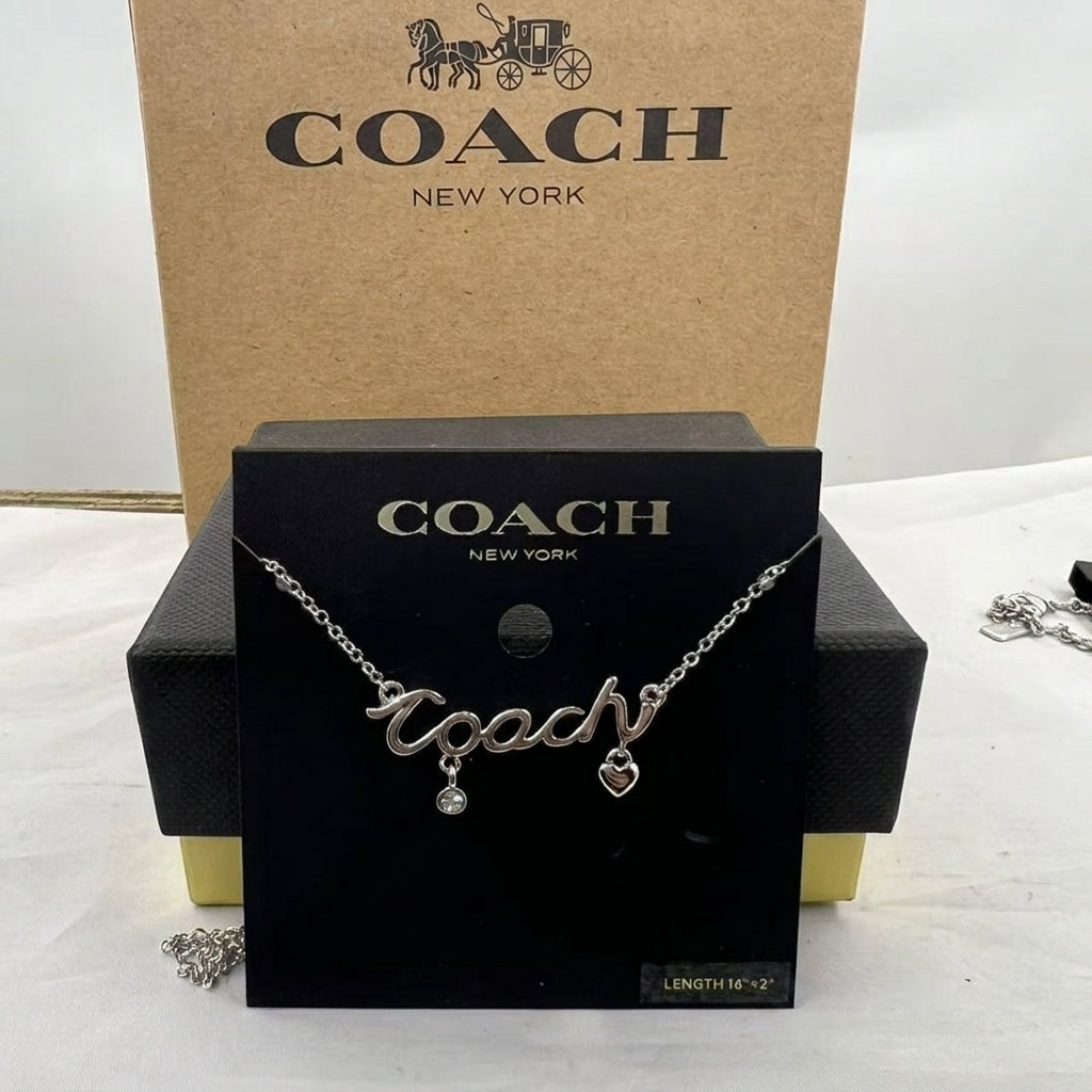 A silver-tone Coach nameplate necklace with a dangling heart charm and clear rhinestone, displayed on a black Coach box against a brown Coach New York box.