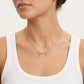 A close-up of a model wearing a silver script nameplate necklace that reads 'Coach' with small dangling charms, paired with a white ribbed tank top against a light neutral background.