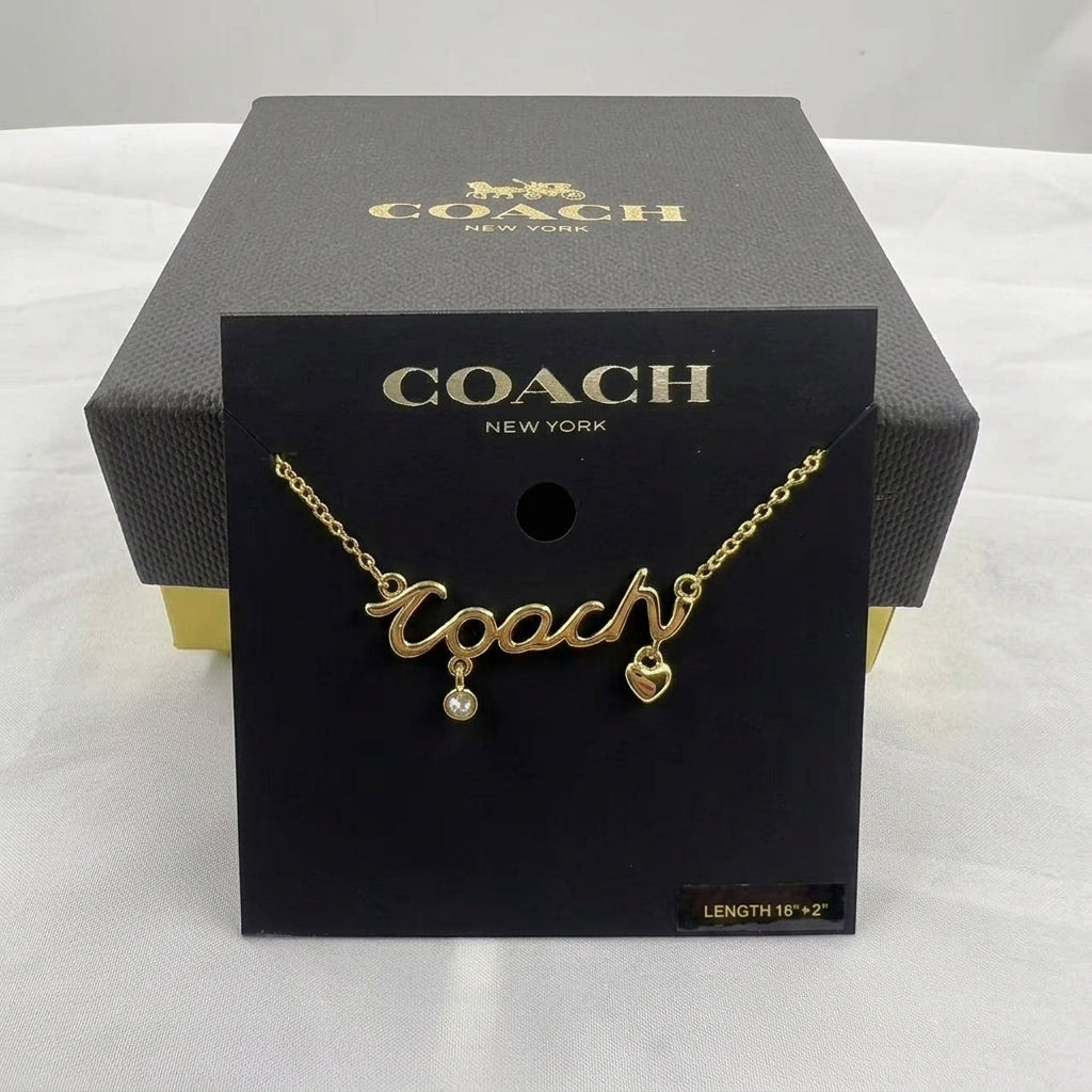 A gold-tone Coach necklace with a cursive 'Coach' pendant, a small dangling pearl, and a heart charm, displayed on a black Coach New York card insert in front of its black branded box.