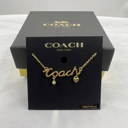 A gold-tone Coach necklace with a cursive 'Coach' pendant, a small dangling pearl, and a heart charm, displayed on a black Coach New York card insert in front of its black branded box.