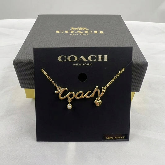 A gold-tone Coach necklace with a cursive 'Coach' pendant, a small dangling pearl, and a heart charm, displayed on a black Coach New York card insert in front of its black branded box.