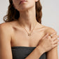 A close-up of a woman wearing a delicate gold necklace with a crescent moon pendant and a gold ring on her finger, styled with a dark gray strapless top against a light gray background.