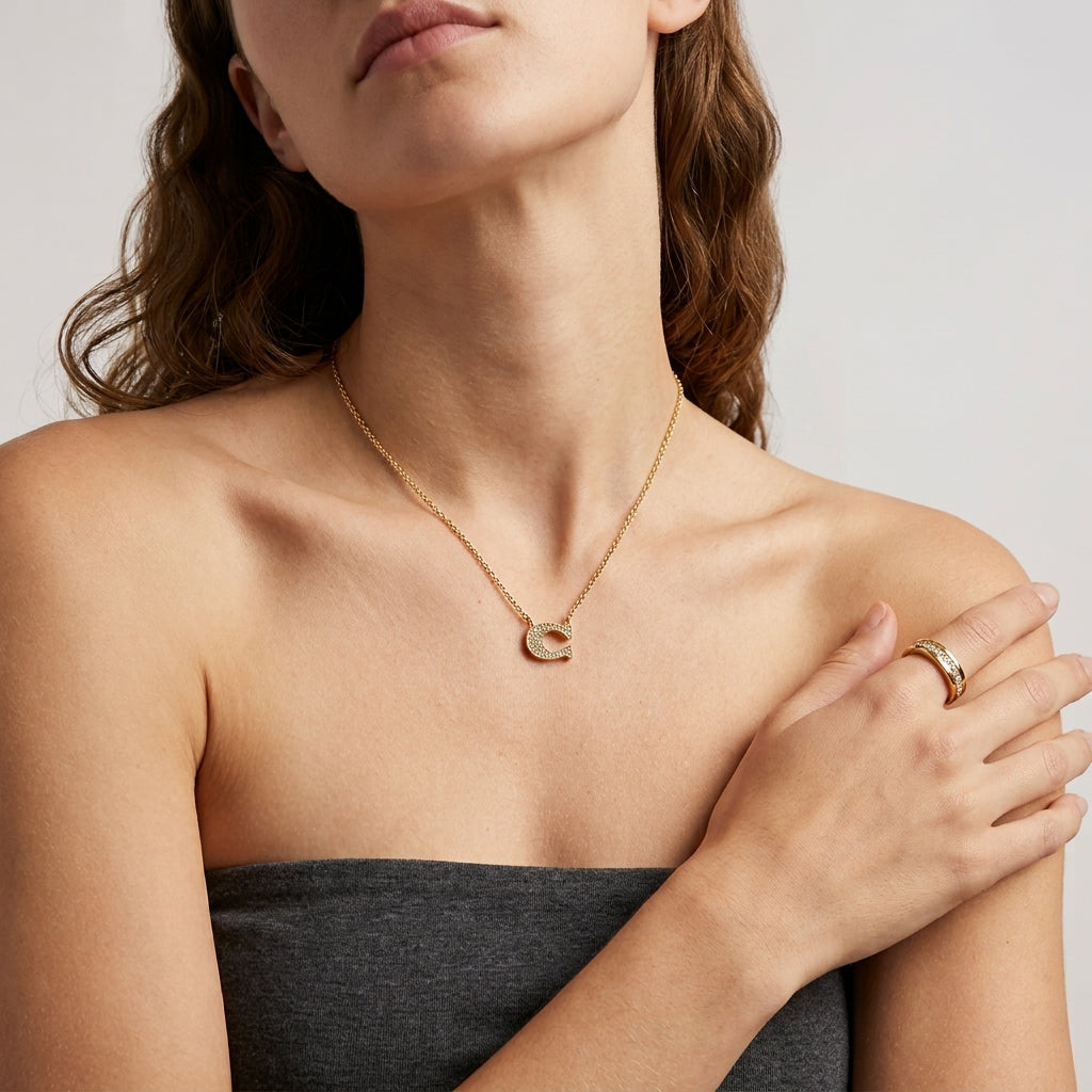 A close-up of a woman wearing a delicate gold necklace with a crescent moon pendant and a gold ring on her finger, styled with a dark gray strapless top against a light gray background.