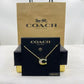 A Coach New York necklace displayed on a black branded card, with a kraft Coach shopping bag behind it. The necklace has a gold-tone chain and a letter C pendant set with iridescent stones.
