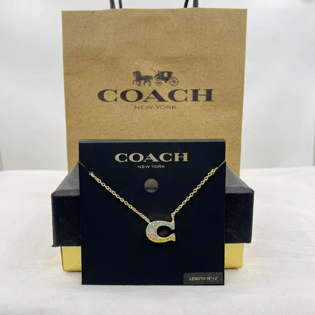 A Coach New York necklace displayed on a black branded card, with a kraft Coach shopping bag behind it. The necklace has a gold-tone chain and a letter C pendant set with iridescent stones.