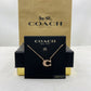 A Coach letter C initial necklace in rose gold with a textured pavé center, displayed on a black branded card holder with a gold and black Coach box behind it.