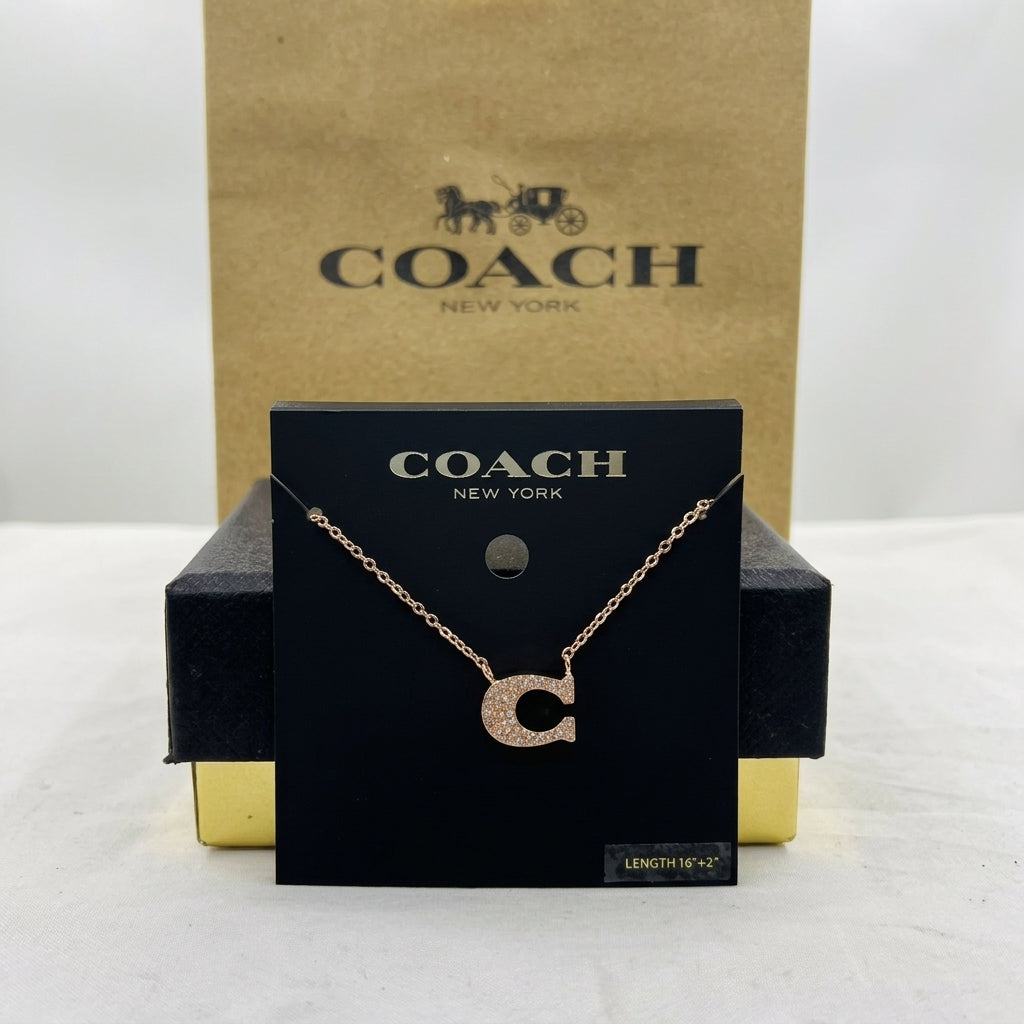 A Coach letter C initial necklace in rose gold with a textured pavé center, displayed on a black branded card holder with a gold and black Coach box behind it.