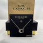 A silver initial necklace with a sparkling letter 'C' pendant, displayed on a black Coach New York card, with its brown and gold packaging box behind it.