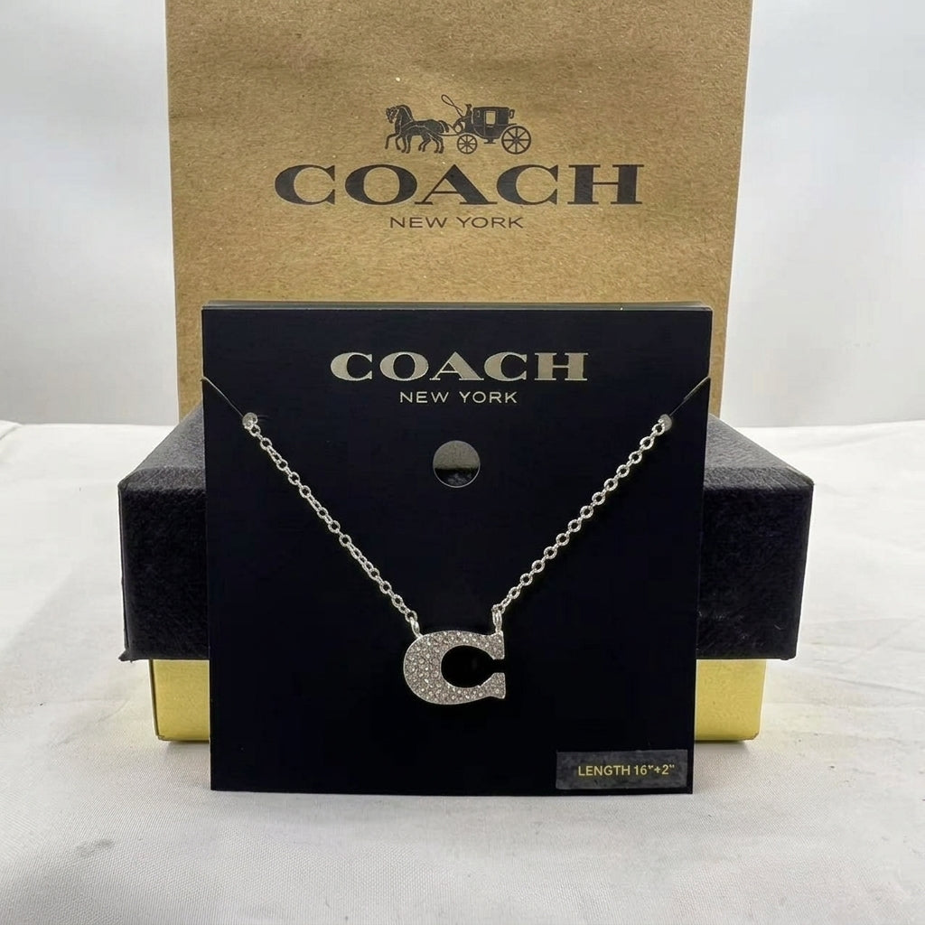 A silver initial necklace with a sparkling letter 'C' pendant, displayed on a black Coach New York card, with its brown and gold packaging box behind it.