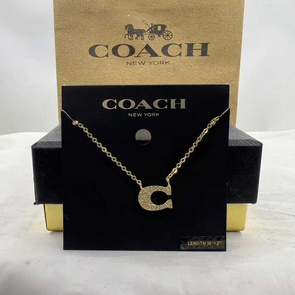 A gold initial necklace with a sparkling letter 'C' pendant, displayed on a black Coach New York card hanger in front of a brown Coach New York box.