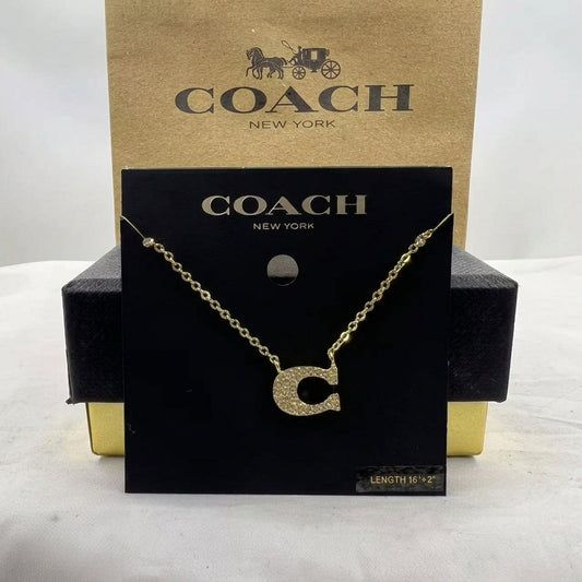 A gold initial necklace with a sparkling letter 'C' pendant, displayed on a black Coach New York card hanger in front of a brown Coach New York box.