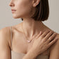 A close-up lifestyle shot of a woman wearing a delicate gold chain necklace with a small pink butterfly pendant, styled with gold rings and hoop earrings against a neutral beige background.