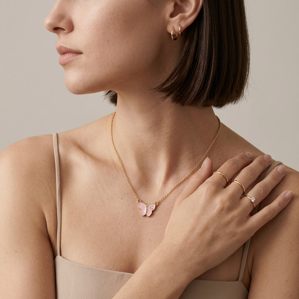A close-up lifestyle shot of a woman wearing a delicate gold chain necklace with a small pink butterfly pendant, styled with gold rings and hoop earrings against a neutral beige background.