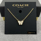 A gold-tone Coach New York butterfly necklace displayed on a black branded card, with the chain draped across the front and a small size label reading 16+2 inches.