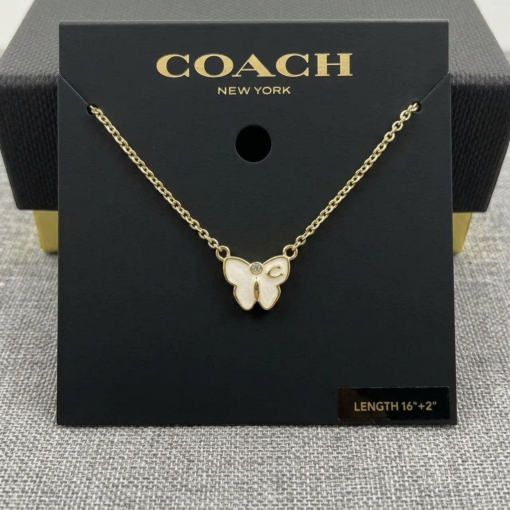 A gold-tone Coach New York butterfly necklace displayed on a black branded card, with the chain draped across the front and a small size label reading 16+2 inches.