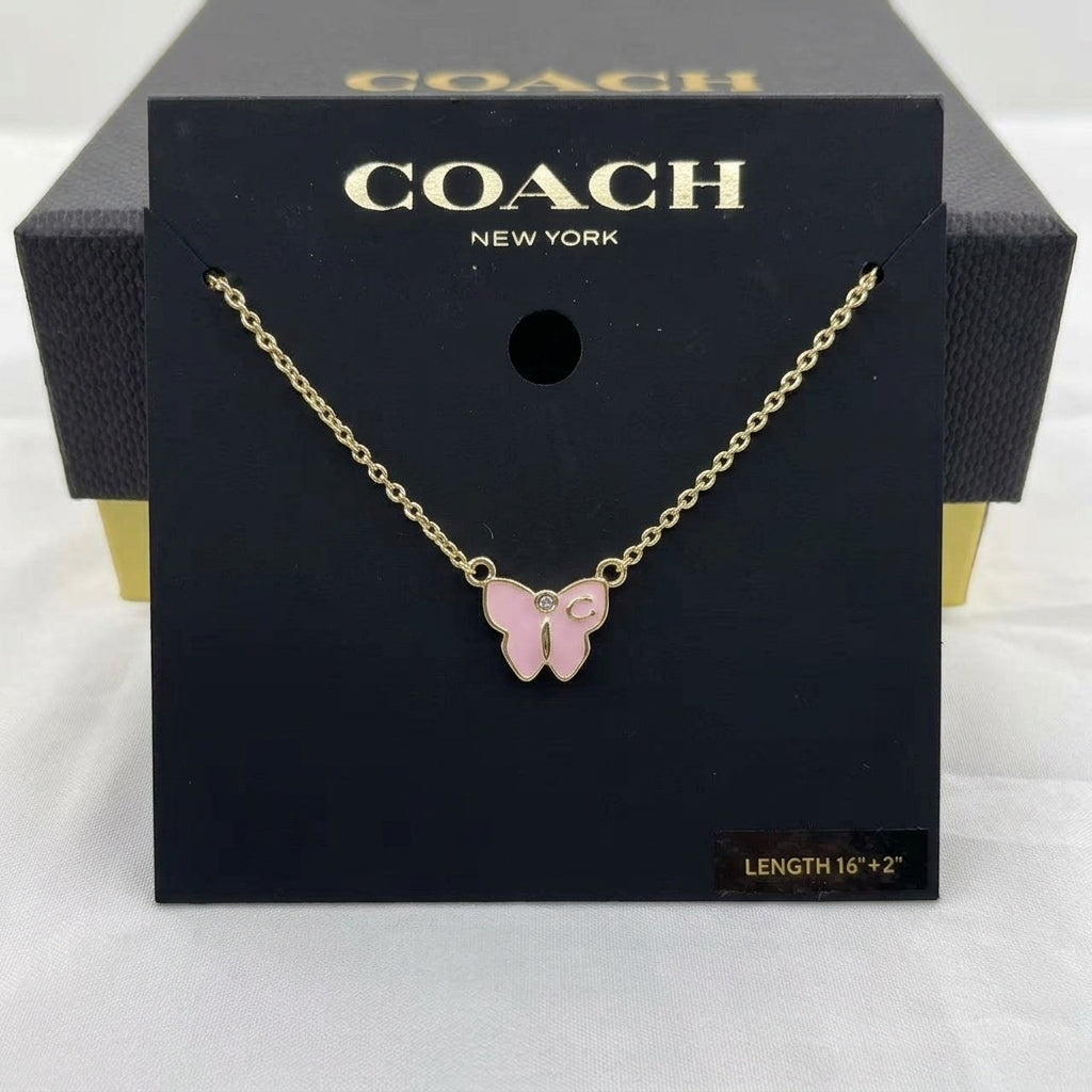 A Coach New York necklace with a pink butterfly pendant on a gold chain, displayed on its black branded packaging card.
