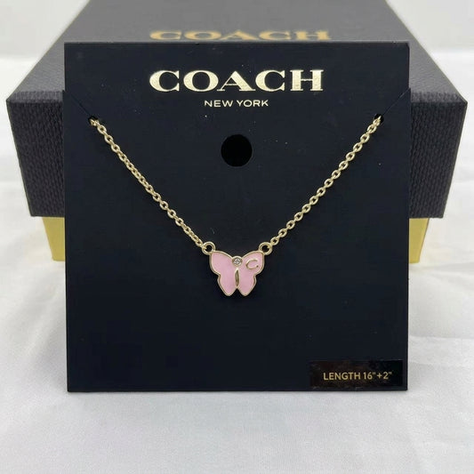 A Coach New York necklace with a pink butterfly pendant on a gold chain, displayed on its black branded packaging card.