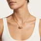 A close-up of a model wearing a delicate gold chain necklace with a small pendant featuring pink enamel and gold details, paired with a white ribbed tank top against a light beige background.